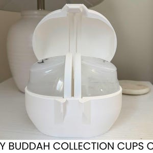 May include: A white plastic case with an open lid, designed to hold baby cups. The case has a rounded shape with a handle and the text "BABY BUDDAH COLLECTION CUPS CASE" at the bottom. The case is sitting on a white surface.