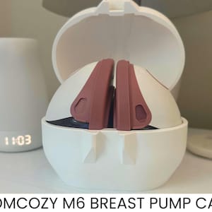 以下が含まれることがあります： 「MOMCOZY M6 BREAST PUMP CASE」の文字が入った白とモーヴ色の搾乳器ケース。ケースは開いており、ポンプのコンポーネントが見えます。左側には、11:03と表示されたデジタル時計が付いた白い円筒形の物体があります。