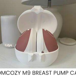May include: A white Momcozy M9 breast pump case, open to show two pink and white breast pump components. The case is on a white surface, with a white lamp and a small digital clock displaying 11:54 in the background.