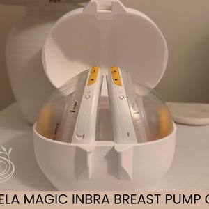 Puede incluir: Un estuche blanco Medela Magic Inbra para extractor de leche, abierto para revelar dos componentes del extractor. Los componentes son blancos y transparentes con detalles amarillos. El estuche está sobre una superficie blanca.