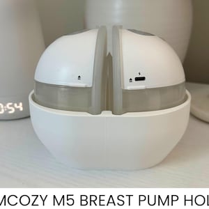 May include: White breast pump holder with a rounded design. The holder is split into two sections, with a clear, cylindrical container at the base. The product has a charging port and an arrow symbol. The text "MOMCOZY M5 BREAST PUMP HOLDER" is at the bottom.