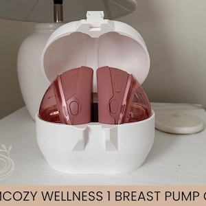 May include: A white breast pump case with a hinged lid, open to reveal two pink breast pumps. The case is on a white surface, with a white lamp and a small, round object in the background. The text "MOMCOZY WELLNESS 1 BREAST PUMP CASE" is at the bottom.