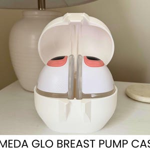 May include: White Ameda Glo breast pump case, open to reveal two white breast pump components with pink accents. The case is on a white surface, with a white lamp and a small, round, beige object in the background.