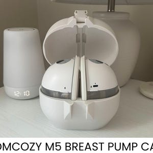 May include: A white Momcozy M5 breast pump case, open to reveal the pump components. A white bedside lamp with a digital clock showing 12:10 is to the left. A white lamp and a small, round, white object are in the background.