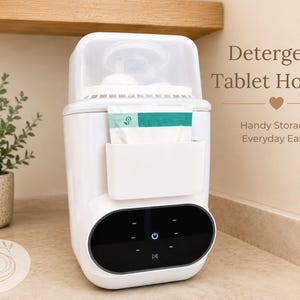 Puede incluir: Un soporte blanco para tabletas de detergente con tapa superior transparente y bolsillo de almacenamiento frontal. El dispositivo cuenta con un panel de control digital negro con icono de encendido y botón de reproducción. Texto: Detergent Tablet Holder.