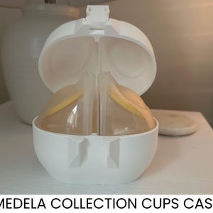 May include: A white Medela Collection Cups Case, open to reveal two clear plastic cups with yellow accents. The case is on a white surface, with the text "MEDELA COLLECTION CUPS CASE" at the bottom.