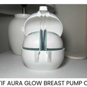 May include: White Motif Aura Glow breast pump case, open to reveal the pump components. The case has a rounded shape with a hinged lid. The pump components are white with teal accents. The text "MOTIF AURA GLOW BREAST PUMP CASE" is at the bottom.