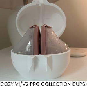 May include: White case holding two breast pump collection cups. The cups are pale pink and enclosed in clear plastic. The case is rounded, and the text "MOMCOZY V1/V2 PRO COLLECTION CUPS CASE" is at the bottom of the image.