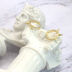 May include: Gold-colored hoop earrings with a spiky design and small, clear stones. The earrings are displayed on a white sculpture with a marble-like background. The earrings have a unique, edgy style.