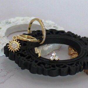 May include: A collection of gold-colored jewelry, including a sun charm hoop earring, a ring with small clear stones, a lightning bolt stud earring, and a cluster stud earring with three clear stones. The jewelry is displayed on a black decorative frame.