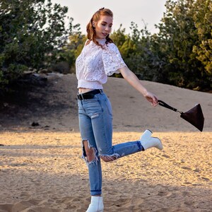 May include: A woman wearing a white lace top, blue jeans with rips, and white ankle boots. She is holding a brown leather purse and is standing in a sandy area.