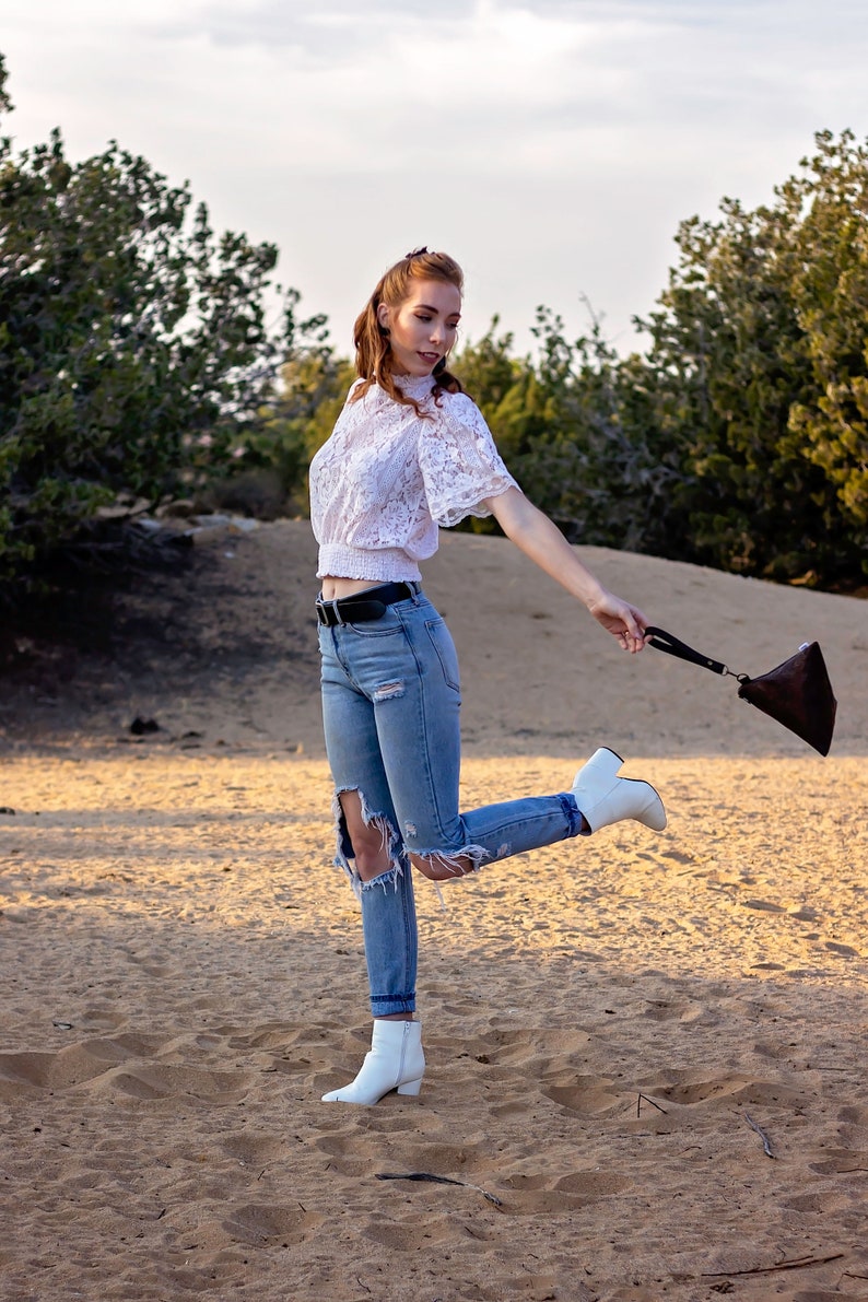 May include: A woman wearing a white lace top, blue jeans with rips, and white ankle boots. She is holding a brown leather purse and is standing in a sandy area.