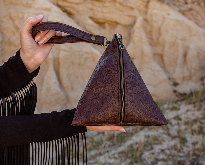 May include: A brown leather triangular purse with a zipper closure and a floral embossed design. The purse has a brown leather strap.