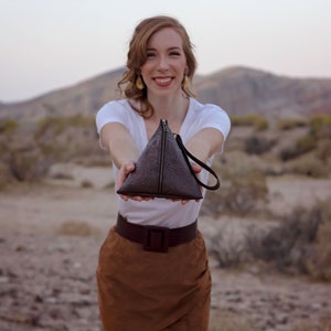 May include: A woman is holding a brown triangular purse with a zipper and a strap. The purse is made of textured leather. The woman is wearing a white t-shirt and a brown skirt.