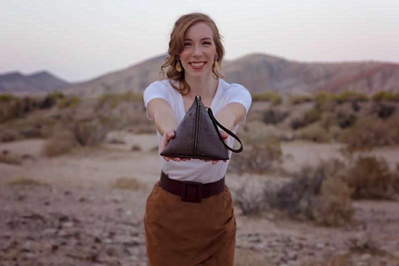 May include: A woman is holding a brown triangular purse with a zipper and a strap. The purse is made of textured leather. The woman is wearing a white t-shirt and a brown skirt.