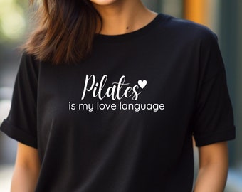 Peut inclure: T-shirt noir avec un texte blanc qui dit "Pilates is my love language" et un petit cœur blanc.
