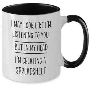 May include: White coffee mug with a black rim and handle. The mug has black text that reads "I may look like I'm listening to you but in my head I'm creating a spreadsheet."