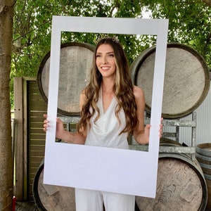 Può includere: Una donna indossa un top bianco con paillettes e una fascia dorata con la scritta "Bride to Be" e tiene un grande telaio bianco per foto booth davanti a sé. Il telaio è davanti a una pila di botti di legno.