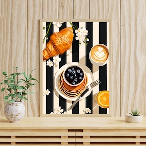 Pu&ograve; includere: Una stampa artistica da parete incorniciata che raffigura una scena di colazione con un croissant dorato, una pila di pancake guarniti con mirtilli in una ciotolina, una tazza di caff&egrave; latte art, un bicchiere di succo d'arancia e fiori bianchi su sfondo a righe bianche e nere.