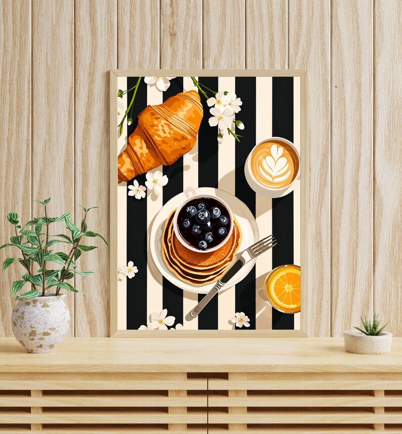 Pu&ograve; includere: Una stampa artistica da parete incorniciata che raffigura una scena di colazione con un croissant dorato, una pila di pancake guarniti con mirtilli in una ciotolina, una tazza di caff&egrave; latte art, un bicchiere di succo d'arancia e fiori bianchi su sfondo a righe bianche e nere.