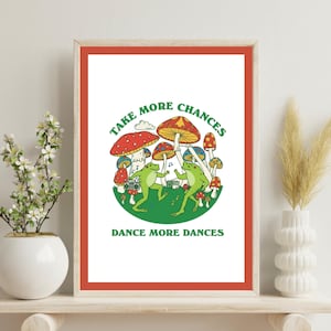 May include: A framed art print with a red border displays a whimsical illustration. Two green frogs dance amongst colourful mushrooms. The text reads "TAKE MORE CHANCES" and "DANCE MORE DANCES" in green. The artwork has a retro, psychedelic vibe.