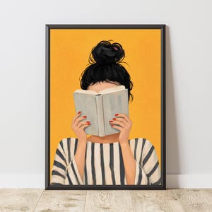 Op de afbeelding: Ingelijste kunstprint van een persoon met zwart haar in een knot, die een boek leest. De handen, met rode nagellak, houden het boek voor het gezicht. Ze draagt een zwart-wit gestreept shirt tegen een gele achtergrond.