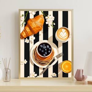 Pu&ograve; includere: Una stampa artistica incorniciata che raffigura una scena di colazione su uno sfondo a righe bianche e nere. L'immagine mostra un croissant dorato, una pila di pancake con mirtilli, una tazza di caff&egrave; con latte art e succo d'arancia.