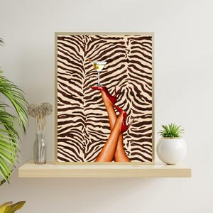 May include: A framed print featuring a martini glass balanced on the feet of a person wearing red high heels. The background is a zebra print pattern in shades of brown and beige. The artwork is displayed on a wooden shelf.