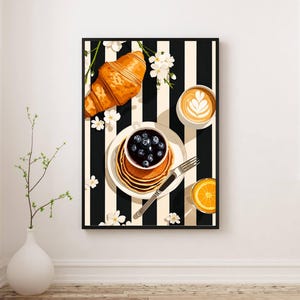 Pu&ograve; includere: Una stampa d'arte incorniciata che raffigura una scena di colazione su uno sfondo a righe bianche e nere. L'immagine mostra un croissant dorato, una pila di pancake guarniti con mirtilli, una tazza con latte art e un bicchiere di spremuta.