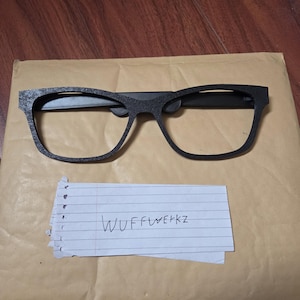 May include: Black-framed eyeglasses with a rectangular shape, resting on a brown envelope. The text "WUFFWERKZ" is handwritten on a piece of paper beneath the glasses. The glasses have a matte finish.