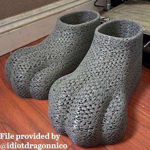 May include: Two gray, 3D-printed foot-shaped objects with a textured, open-weave design. The objects have a claw-like appearance and are displayed on a wooden surface. The text "File provided by @idiotdragonnico" is visible at the bottom.