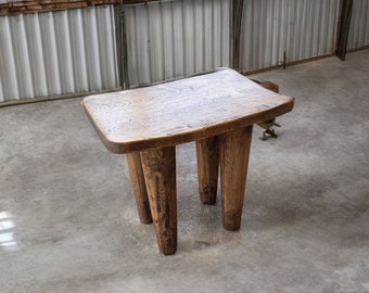Puede incluir: Un taburete de madera con una tapa rectangular y cuatro patas. El taburete está hecho de madera oscura con un acabado natural. El taburete está sentado en un suelo de hormigón.