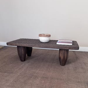 Op de afbeelding: Een donkerbruine houten salontafel met vier dikke, afgeronde poten. De tafel heeft een ruw, getextureerd oppervlak en is versierd met een witte keramische kom en een boek.