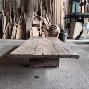 Könnte beinhalten: Ein rustikaler Holz-Couchtisch mit natürlicher Holzoptik. Der Tisch hat ein schlichtes Design mit einer rechteckigen Tischplatte und einer einzigen, dicken Basis. Der Tisch steht auf einem Betonboden.