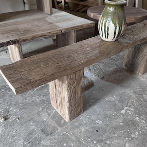 May include: A rustic wooden bench with a green and white glazed ceramic vase on top. The bench is made of rough-hewn wood with a natural finish. The vase has a rounded shape and a simple design.