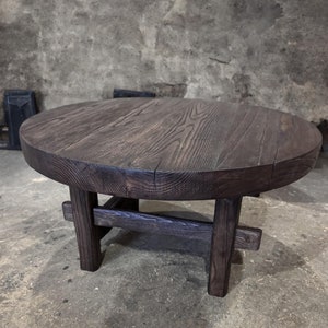 May include: A dark brown round wooden coffee table with a simple, rustic design. The table has a thick, solid top and three legs that are connected by a crossbar.