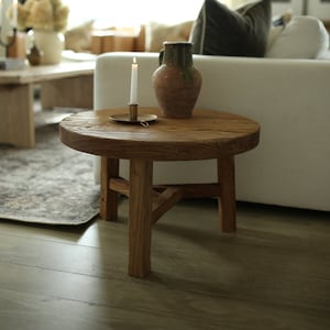 Pode incluir: Uma mesa de apoio redonda de madeira com acabamento rústico e um design simples e robusto. A mesa possui um único porta-velas e um jarro de cerâmica.