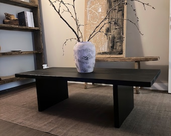May include: A black wooden coffee table with a unique design featuring two large rectangular legs on either side. The table is placed on a brown rug in a room with a white vase filled with dried branches and a black and white abstract painting on the wall.