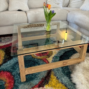 May include: A clear glass-top coffee table with a light wood frame. A vase of tulips sits on the table. The table is in front of a cream-colored sectional sofa with decorative pillows. A colorful rug is beneath the table.