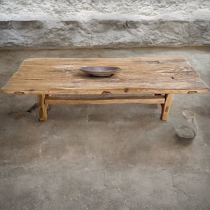 May include: A rustic, rectangular wooden coffee table with a natural finish. The table features a distressed look, with visible wood grain and knots. A small, round, ceramic bowl sits on the table's surface. A clear glass vase is on the floor.