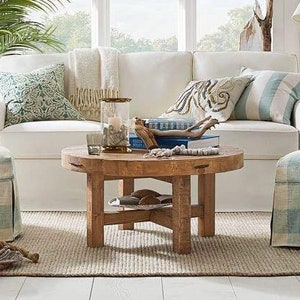 Peut inclure: Un canapé blanc avec des coussins à motifs bleus et blancs, une table basse en bois et un pouf en tissu bleu et blanc à carreaux dans un salon avec une grande fenêtre et une plante en pot.