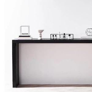 May include: A black console table with minimalist decor. The table holds a Newton's cradle, a white ceramic bowl, a black tray with bottles, a geometric metal sculpture, a stack of books, and a dark glass bottle with branches.