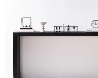 Puede incluir: Una consola negra con decoración minimalista. La mesa contiene una cuna de Newton, un cuenco de cerámica blanco, una bandeja negra con botellas, una escultura geométrica de metal, una pila de libros y una botella de vidrio oscuro con ramas.