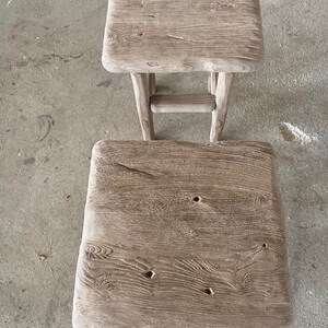 May include: Two rustic wooden stools with a weathered gray finish. The top stool has a simple rectangular shape, while the bottom stool has a slightly rounded top. Both stools have a natural wood grain pattern and show signs of age and use.