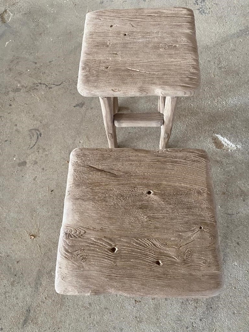 May include: Two rustic wooden stools with a weathered gray finish. The top stool has a simple rectangular shape, while the bottom stool has a slightly rounded top. Both stools have a natural wood grain pattern and show signs of age and use.