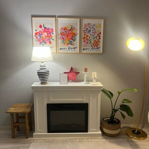 May include: A white fireplace mantel with a black electric fireplace insert. The mantel has a wooden stool in front of it and a potted plant to the right. Three framed floral prints hang on the wall above the mantel. The prints feature colorful flowers and the text "Bloom Basket London" and "Bloom Basket Hokkaido".