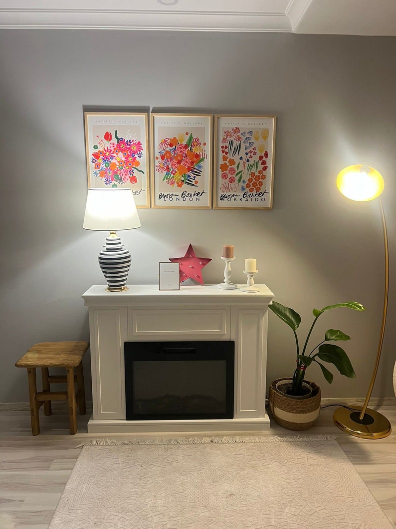 May include: A white fireplace mantel with a black electric fireplace insert. The mantel has a wooden stool in front of it and a potted plant to the right. Three framed floral prints hang on the wall above the mantel. The prints feature colorful flowers and the text "Bloom Basket London" and "Bloom Basket Hokkaido".