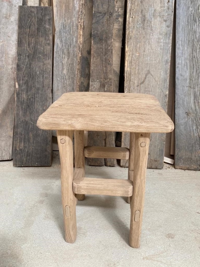 May include: A rustic wooden stool with a square top and a lower shelf. The stool is made of natural wood with a light brown finish. It has a simple design with rounded edges and a sturdy construction.