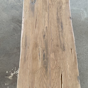 May include: A weathered, rectangular wooden bench with a natural, light brown finish. The bench features visible wood grain, knots, and cracks, showcasing its rustic character. The design is simple and sturdy, suitable for various settings.