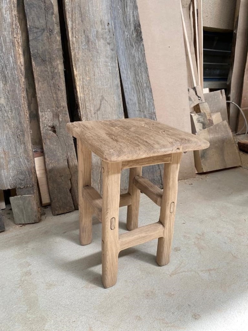 May include: A rustic wooden stool with a square top and four legs. The stool is made of natural wood with a light brown finish. The legs are slightly curved and have a simple design.